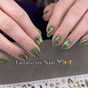 May include: Green nail art with gold accents and fantasy-themed designs, including moons, stars, and butterflies. The text "Fantasycore Nails" is displayed below the nails. The nails are oval-shaped.