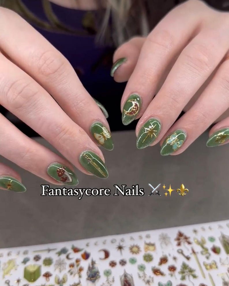 May include: Green nail art with gold accents and fantasy-themed designs, including moons, stars, and butterflies. The text "Fantasycore Nails" is displayed below the nails. The nails are oval-shaped.