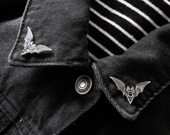 May include: Two silver bat pins on a black denim jacket with a white and black striped fabric underneath.