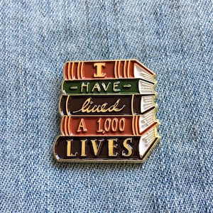 Può includere: Una spilla smaltata colorata a forma di pila di libri. I libri sono in tonalità di rosso, verde e marrone, con scritte dorate che recitano "I HAVE lived A 1,000 LIVES". La spilla ha un bordo dorato.