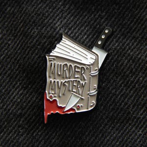 May include: A silver-colored enamel pin in the shape of a book with the words "MURDER MYSTERY" on the cover. A knife is piercing the book, with a red blood stain dripping down. The pin is on a dark gray fabric.