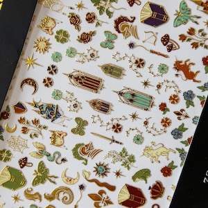 May include: Sheet of decorative stickers featuring various gold foil designs. Includes butterflies, flowers, stars, and architectural elements in shades of green, blue, and red. The stickers have a metallic gold outline and are arranged on a white background.