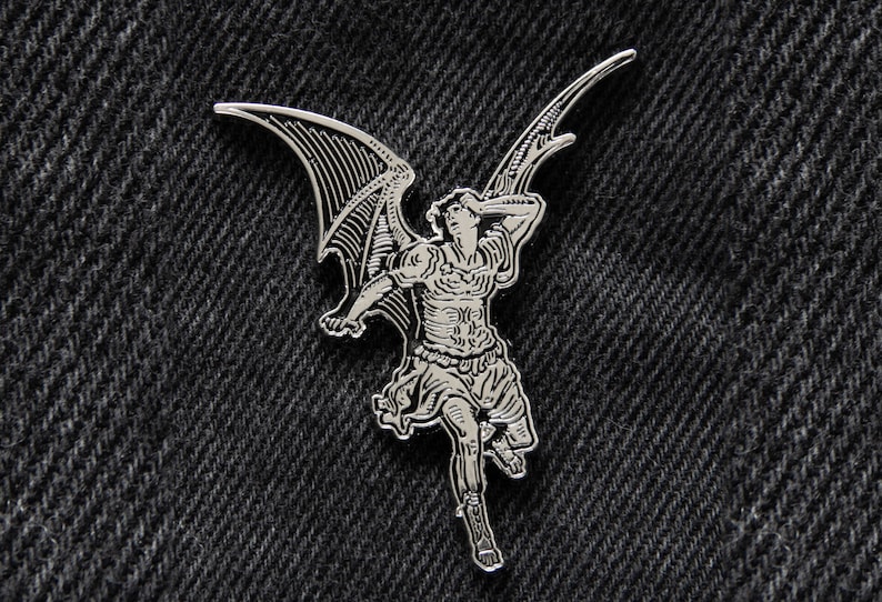 May include: A silver-toned metal pin featuring a detailed illustration of an angel with wings outstretched. The angel is depicted in a dynamic pose, with its wings spread wide and its body leaning forward.