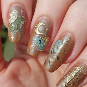 May include: Close-up of manicured nails with a shimmering brown polish. Each nail features gold and teal embellishments, including a frog with wings, a crescent moon, butterflies, stars, and a spiderweb. The nails are long and oval-shaped.