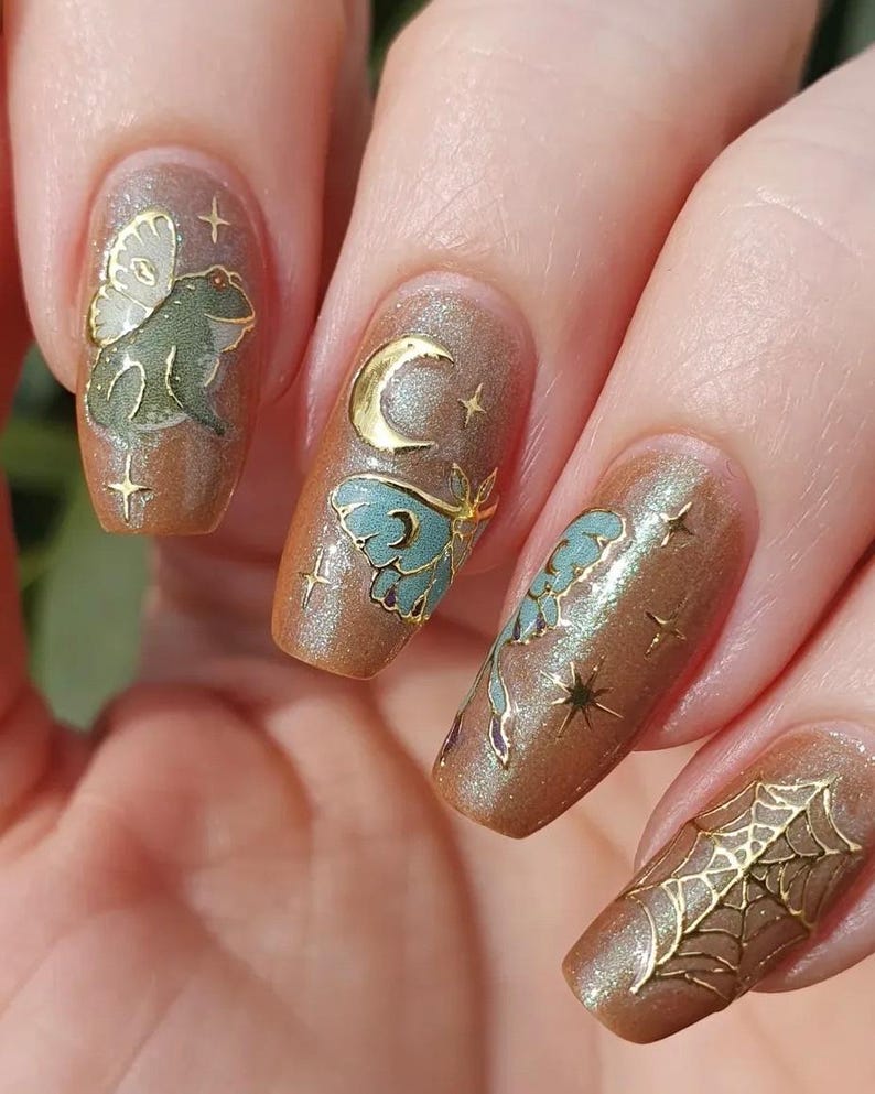 May include: Close-up of manicured nails with a shimmering brown polish. Each nail features gold and teal embellishments, including a frog with wings, a crescent moon, butterflies, stars, and a spiderweb. The nails are long and oval-shaped.