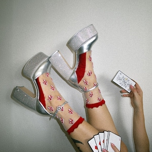 May include: A pair of silver platform shoes with rhinestones, worn with red sheer socks with mushroom designs. The socks have a red ruffle trim. The person is holding a deck of playing cards.