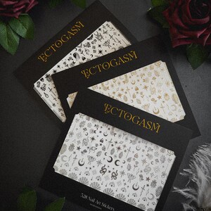 K&ouml;nnte beinhalten: Vier schwarze Kartonverpackungen mit Nagelkunst-Aufklebern mit goldenem Text "ECTOGASM". Die Aufkleber zeigen verschiedene Designs, darunter Monde, Sterne, Schlangen und Schmetterlinge. Die untere Verpackung tr&auml;gt die Aufschrift "528 Nail Art Stickers".