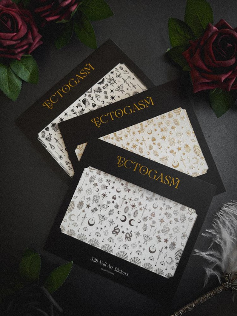 K&ouml;nnte beinhalten: Vier schwarze Kartonverpackungen mit Nagelkunst-Aufklebern mit goldenem Text "ECTOGASM". Die Aufkleber zeigen verschiedene Designs, darunter Monde, Sterne, Schlangen und Schmetterlinge. Die untere Verpackung tr&auml;gt die Aufschrift "528 Nail Art Stickers".
