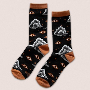 May include: A pair of black socks featuring a repeating pattern of stylized eyes, clouds, and stars. The socks have brown heels, toes, and cuffs. The design incorporates white, grey, and brown elements.