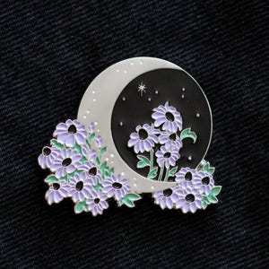 May include: A decorative enamel pin featuring a crescent moon with a black interior and a starry night design. Lavender flowers with green leaves are clustered around the moon. The pin has a silver border and is set against a dark background.