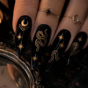 K&ouml;nnte beinhalten: Schwarze, lange, spitze Fingern&auml;gel mit goldenen Himmelsdesigns, darunter Monde, Sterne und Schlangen. Die N&auml;gel sind mit goldenen Ringen verziert. Eine brennende Kerze und ein dekorativer Spiegel befinden sich im Hintergrund.