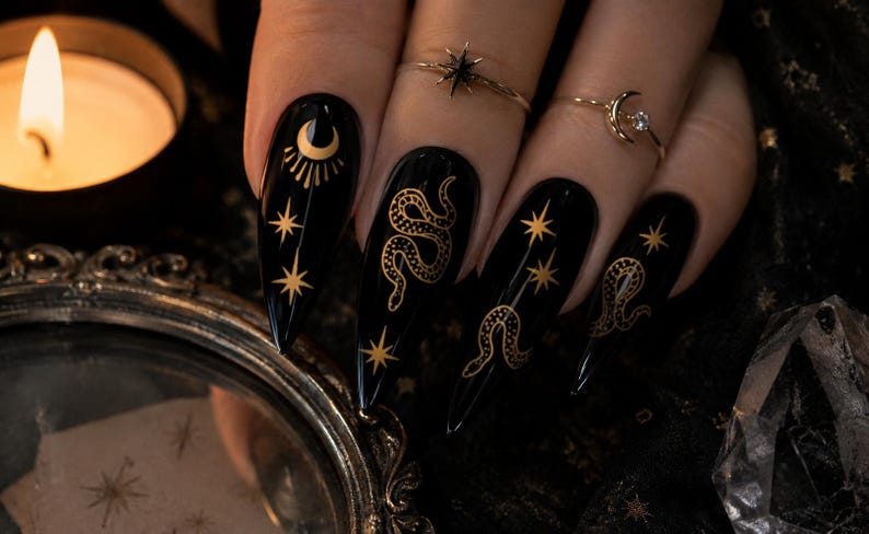 K&ouml;nnte beinhalten: Schwarze, lange, spitze Fingern&auml;gel mit goldenen Himmelsdesigns, darunter Monde, Sterne und Schlangen. Die N&auml;gel sind mit goldenen Ringen verziert. Eine brennende Kerze und ein dekorativer Spiegel befinden sich im Hintergrund.