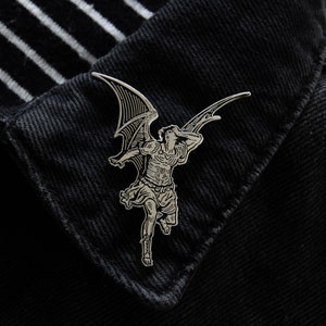 May include: Silver-toned metal pin featuring a detailed illustration of an angel with wings outstretched. The angel is depicted in a dramatic pose, with its head thrown back and its wings spread wide.