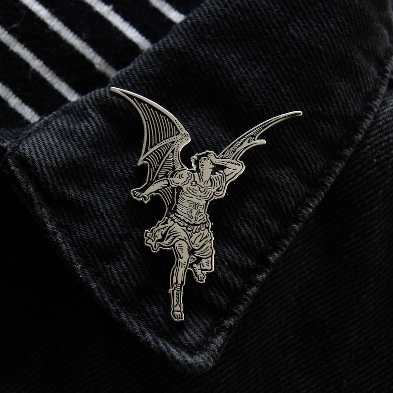May include: Silver-toned metal pin featuring a detailed illustration of an angel with wings outstretched. The angel is depicted in a dramatic pose, with its head thrown back and its wings spread wide.