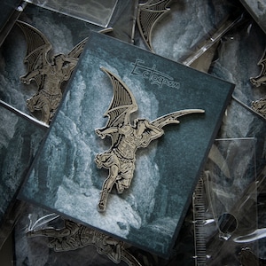 May include: A silver-toned metal pin featuring a detailed illustration of an angel with wings spread wide. The angel is depicted in flight, with its head turned back to look at something behind it. The pin is set against a dark blue background with a white, textured design. The text "Ectoplasm" is printed in white on the background.