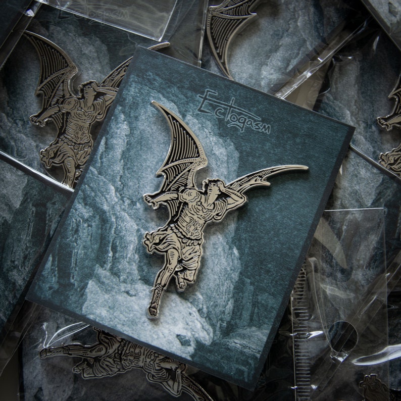 May include: A silver-toned metal pin featuring a detailed illustration of an angel with wings spread wide. The angel is depicted in flight, with its head turned back to look at something behind it. The pin is set against a dark blue background with a white, textured design. The text "Ectoplasm" is printed in white on the background.