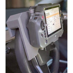 May include: A grey 3D-printed mount attached to a black steering wheel, holding a dark grey DJI drone remote controller with a built-in screen displaying flight data. The controller features dual joysticks, various buttons, and a metallic knob.