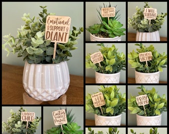 May include: A collage of 10 images featuring potted plants with humorous wooden signs. The signs have various sayings, including "Emotional Support Plant", "What the Fucculent", "I Will Survive", "Water Please", "My Next Victim", "Favorite Child", "Cactus Makes Perfect", "Feeling Cute Might Die Later", "Pray For Me", "Fake", "Please Don't Die", "Unmotivated But Willing To Pretend I'm Plants", and "I Just Pretend I'm Plants".