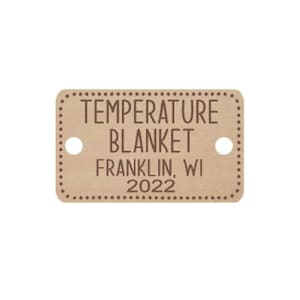 May include: A brown wooden tag with a dotted border and rounded corners. The tag says "TEMPERATURE BLANKET FRANKLIN, WI 2022".