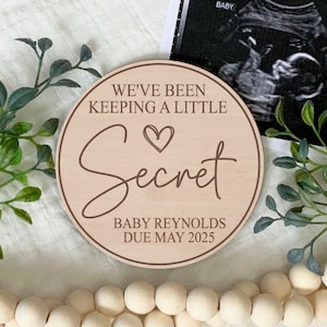 May include: A round wooden sign with the text "We've been keeping a little secret" with a heart symbol and the text "Baby Reynolds Due May 2025".