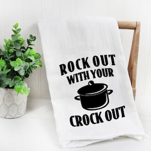 May include: White kitchen towel with the text "ROCK OUT WITH YOUR CROCK OUT" in black. A black illustration of a slow cooker is between the text. The towel is draped over a wooden chair.