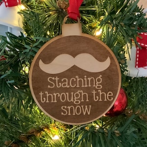 May include: A wooden Christmas ornament shaped like a bauble with a brown background and white text. The ornament has a white mustache and the text "Staching through the snow".