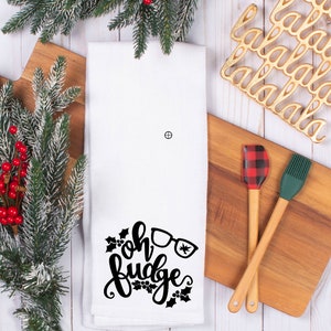 May include: White kitchen towel with black text that reads "Oh Fudge" with a pair of glasses and holly sprigs. The towel is on a white wooden surface with a green and red spatula, a green silicone brush, and a gold metal "la la la" trivet.