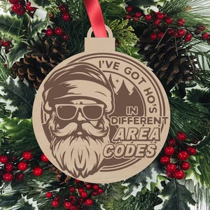 May include: Wooden Christmas ornament with a Santa Claus design wearing sunglasses and a long beard. The ornament has the text "I've got ho's in different area codes" in a circular design. A red ribbon is attached for hanging.