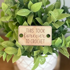 May include: A wooden tag with the text "This took forever to crochet" in brown lettering. The tag is surrounded by a dotted border and has two small holes at the top.