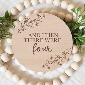 May include: A round wooden sign with the text "And then there were four" in brown lettering. The sign is decorated with a floral design around the edges.