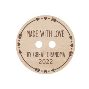 May include: A round wooden button with a brown border and text that reads "Made with Love by Great Grandma 2022". The button has two holes in the center and is decorated with arrows and hearts.