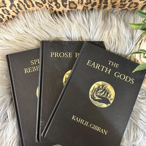 May include: Three black hardcover books with gold lettering. The titles include "Spirit Rebels", "Prose Poems", and "The Earth Gods" by Kahlil Gibran. The books are arranged on a fluffy, light-colored surface.