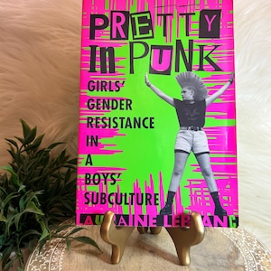 May include: A pink and green book titled "Pretty in Punk: Girls' Gender Resistance in a Boys' Subculture" by Lauraine Leblanc. The cover features a black and white photo of a woman with a mohawk hairstyle wearing a black tank top, denim shorts, and black boots.