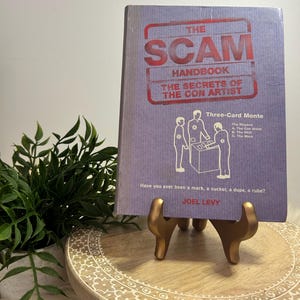 May include: A purple book titled "The Scam Handbook: The Secrets of the Con Artist" by Joel Levy. The book cover features a cartoon illustration of three people playing a three-card monte game.