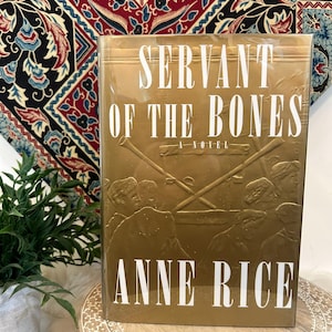 Peut inclure: Un livre doré intitulé "Servant of the Bones" d'Anne Rice. La couverture présente des images en relief et des lettres blanches. Le livre est enveloppé dans du plastique transparent et repose sur un support en bois, avec un tissu à motifs en arrière-plan.