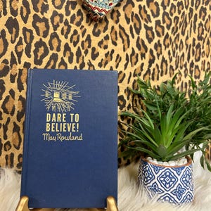 Pode incluir: Um livro azul marinho com letras douradas que diz "DARE TO BELIEVE! May Rowland" é exibido em um suporte dourado. À direita, há uma pequena planta suculenta em vaso com um padrão azul e branco. O fundo apresenta uma estampa de leopardo e um tecido estampado.