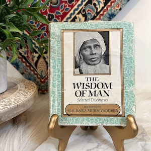 Puede incluir: Un libro antiguo titulado "The Wisdom of Man" con un retrato en blanco y negro de un hombre en la portada. El libro se exhibe en un soporte dorado, sobre un fondo de tela estampada y vegetación.