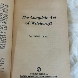 May include: An open book with a tan cover featuring the title "The Complete Art of Witchcraft" by Sybil Leek. The book is a Signet Book from New American Library, Times Mirror.
