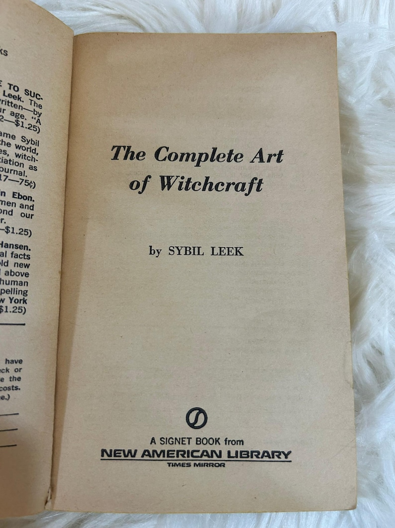 May include: An open book with a tan cover featuring the title "The Complete Art of Witchcraft" by Sybil Leek. The book is a Signet Book from New American Library, Times Mirror.