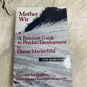 May include: A book titled "Mother Wit: A Feminist Guide to Psychic Development" by Diane Mariechild. The cover features a black and white photo of a sandy beach with a large rock formation in the background. The book is a guide to healing, growth, and spiritual awareness.