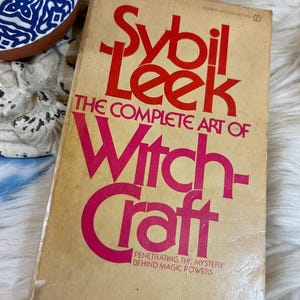 May include: A vintage book titled "Sybil Leek: The Complete Art of Witchcraft" with red and pink lettering on a tan cover. The book's cover has a worn appearance, suggesting its age. The book's subtitle reads "Penetrating the Mystery Behind Magic Powers."