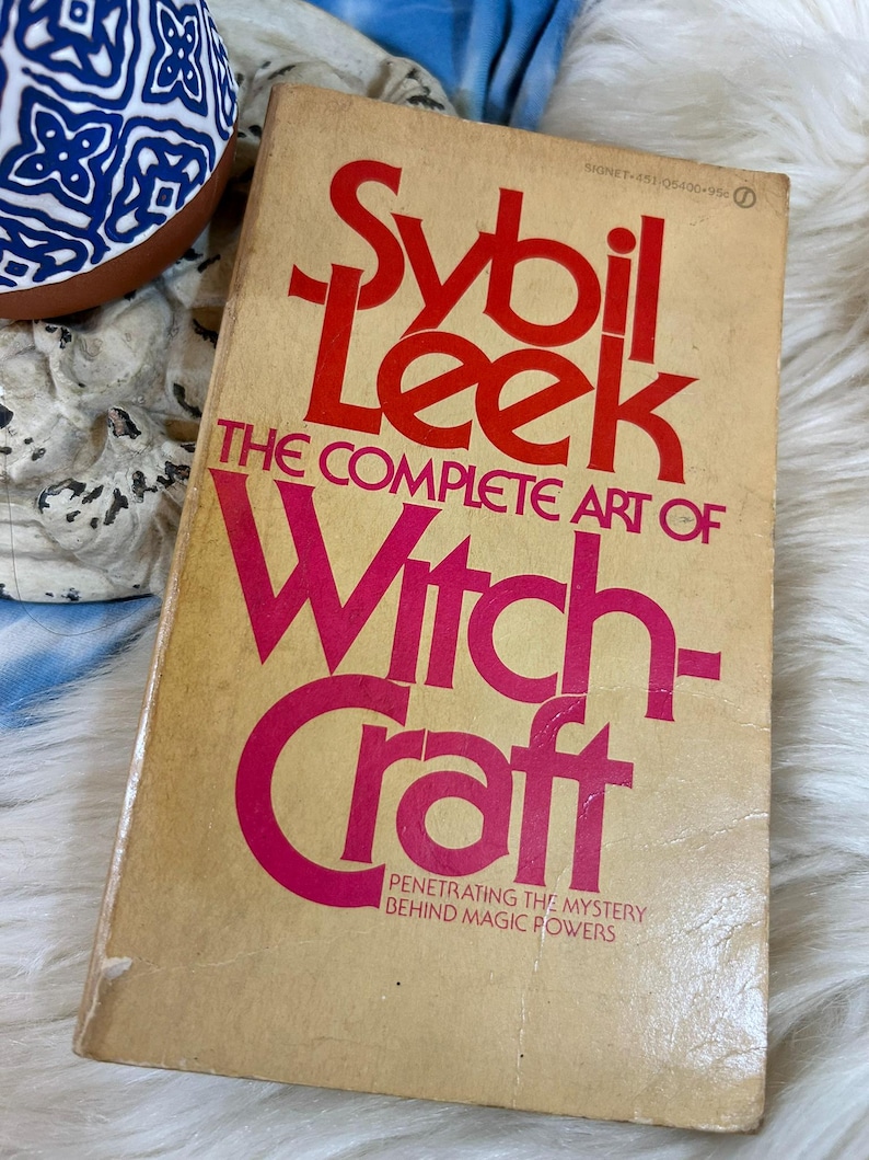 May include: A vintage book titled "Sybil Leek: The Complete Art of Witchcraft" with red and pink lettering on a tan cover. The book's cover has a worn appearance, suggesting its age. The book's subtitle reads "Penetrating the Mystery Behind Magic Powers."