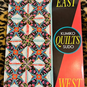May include: A book titled "Kumiko Quilts" by Kumiko Sudo. The cover features a colorful quilt pattern with a black background and the words "East" and "West" on the cover.