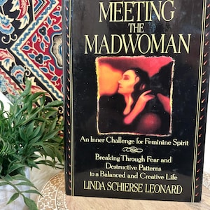 Può includere: Un libro con il titolo "Meeting the Madwoman" con copertina nera e scritte dorate. Il libro presenta un'immagine di una donna sulla copertina. Il libro è su una superficie di legno con uno sfondo a motivi.