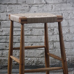 May include: A wooden stool with a woven seat. The stool has a simple design with four legs and a single stretcher.