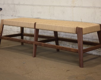 May include: A wooden bench with a woven rope seat and a dark brown finish. The bench has a simple, modern design and is perfect for adding a touch of rustic charm to any space.