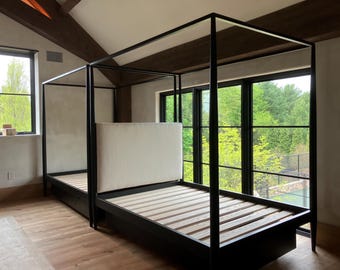 May include: A black metal four-poster bed with a light-colored headboard and wooden slats. The bed frame has a minimalist design and is set against a backdrop of large windows overlooking a green landscape. The room has wooden floors and exposed beams.