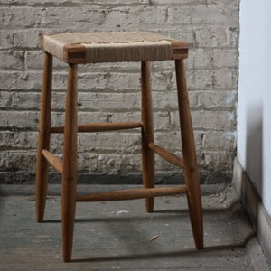 May include: A wooden stool with a woven rope seat. The stool has a simple design with four legs and a rectangular seat.