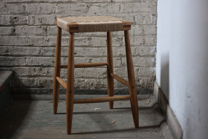 May include: A wooden stool with a woven rope seat. The stool has a simple design with four legs and a rectangular seat.