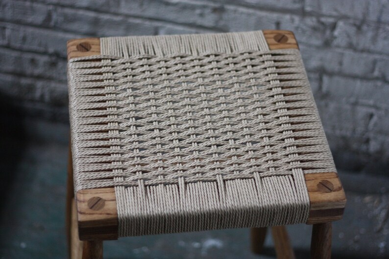 May include: A wooden stool with a woven seat made of natural fiber rope. The seat is woven in a tight, intricate pattern.